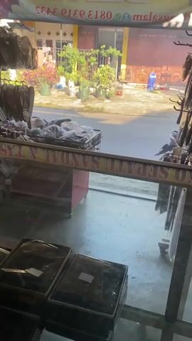 The video shows a street scene viewed from inside a shop. The shop sells shoes, sandals, and bags, as suggested by the writing on the glass window. A display of merchandise lines the top and the bottom of the frame. Outside, a motorcycle with two people riding passes by on a paved road. A house and several potted plants are visible across the street. A person in a blue robe stands near the house. The scene is well-lit, with bright sunlight visible outside the shop.