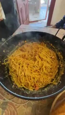 Here is a detailed description of the video:

The video shows someone adding shredded cheese to a large pan of spaghetti. The spaghetti is covered in a reddish-yellow sauce. Steam rises from the pan, indicating the dish is hot. The pan sits on a patterned tabletop. A person in a dark, patterned shirt sprinkles cheese from a package. Another person is partly visible sitting near the table. A partially open door is in the background, leading to a brightly lit area. The room appears to be inside a home or kitchen.
