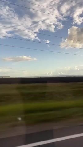 A blurry, low-angle, wide shot captures a view from a moving vehicle, likely a train or car, looking out a window at a landscape. The sky is a pale blue, dotted with fluffy white clouds, some of which are illuminated by sunlight. Thin power lines are visible across the upper portion of the frame, creating horizontal lines. Below the sky, a dark, flat expanse, possibly a field or forest, stretches horizontally. In the foreground, a strip of blurred green and brown represents the passing landscape, with a tree on the right side appearing as a vertical streak of color due to motion blur. The overall impression is one of travel and the fleeting nature of scenery.