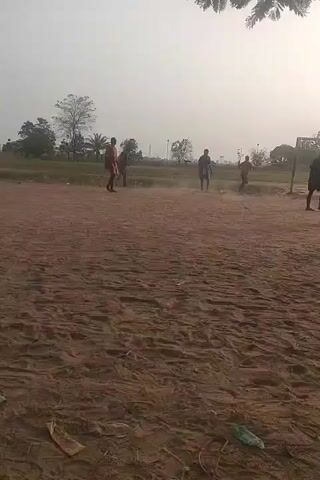 The video shows a group of people playing soccer on a dirt field. There are at least four people visible, and they are all wearing casual clothing. The field is surrounded by trees and grass, and there is a goal at one end. The sky is overcast. The camera is held by someone standing on the field, and it pans slightly to follow the action. The overall tone of the video is casual and informal.