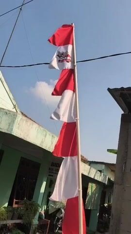 A vertical, low-angle, medium shot shows an Indonesian flag pole against a bright blue sky with some wispy white clouds. The flag is red and white, with an emblem on the top section of the flag. The flag appears to be made of multiple smaller flags tied together, creating a layered effect as it flies in the wind. To the left of the flag pole is a green building with a white sign on it. The sign is mostly unreadable, but some text is visible. To the right of the flag pole, there is a gray wall and a hint of a dark roof. There are also some plants and what appears to be a bicycle visible in the background, partially obscured by the flag pole and building. The overall scene suggests an outdoor setting, possibly in a residential or community area.