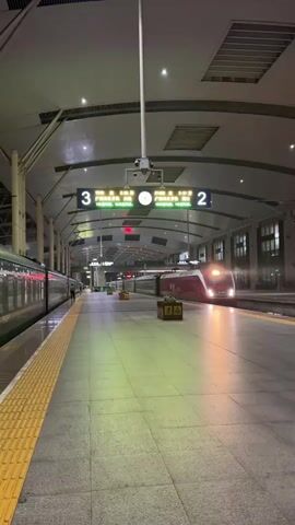 A train station platform is depicted at night. The platform is wide and tiled, with a yellow tactile warning strip along the edge. Two trains are visible, one on the left and a red and white high-speed train on the right. A digital display above the platform shows train information for tracks 3 and 2, including destinations and times. The station has a high, arched roof supported by columns. Artificial lighting illuminates the platform, casting green and yellow hues on the floor. There are no people immediately visible in the foreground, but one person can be seen further down the platform on the left. The overall atmosphere is quiet and still, characteristic of a train station late at night.