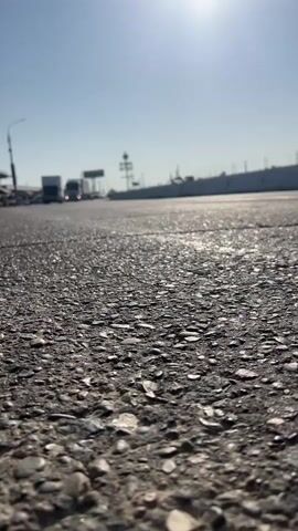 The video shows a low-angle view of an asphalt road with a bright sun in the sky. Several vehicles, including trucks and cars, are driving on the road in the distance. The perspective is close to the ground, focusing on the texture of the asphalt and the tires of passing vehicles.