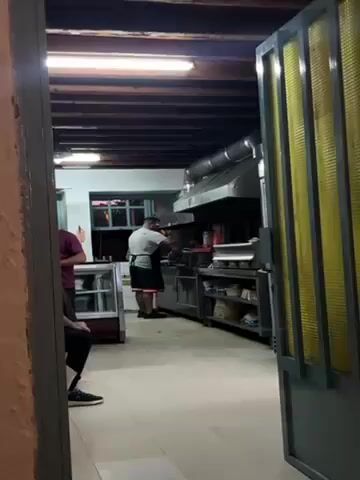 The video shows a glimpse into a restaurant kitchen. A man wearing a white shirt and black apron is behind a stainless steel kitchen counter. The man is facing the cooking station and appears to be cooking or preparing food. The kitchen has a large stainless steel range hood over the cooking area. Several shelves and cabinets are beneath the counter, containing various kitchen supplies. In the foreground, there is an partially open door with a yellow paneled glass. Two people are seen sitting at a table just outside the kitchen. The walls are painted in light colors, and the ceiling has exposed dark wooden beams. A fluorescent light is attached to the ceiling.