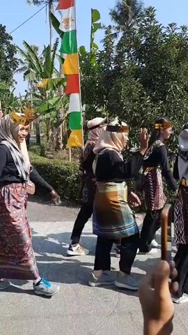 The video shows a group of people walking outdoors along a paved road. Some individuals are wearing Islamic head coverings (hijabs) and traditional clothing. Others are dressed in bright orange shirts and dark pants, with some wearing what appear to be ceremonial headdresses. There are flags and banners visible, including some that resemble the Indonesian flag. Banana trees and other greenery are present in the background, suggesting a tropical or subtropical environment. The sky is clear and blue. The people appear to be participating in a parade or procession, with some waving and smiling. The camera is mostly focused on the individuals as they move forward.