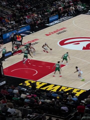 The video captures a high-angle view of a professional basketball game in progress at State Farm Arena, home of the Atlanta Hawks. The court is a light-colored wood with prominent red accents, including a large red hawk logo at mid-court and a solid red painted key area at the end of the court closest to the camera. The baseline features the word "HAWKS" in yellow lettering.

The game is between a team wearing predominantly green uniforms (likely the Boston Celtics, with player numbers visible such as #0, #7, #11, #26, #30) and the home team, the Atlanta Hawks, in white and orange uniforms (players #4, #17, #22 visible).

The action centers around the basket in the foreground. At the 0:00 mark, a player in a green jersey (#0) is in the air, attempting a shot from close range. Several players from both teams are positioned around the key, including a white/orange player (#22) defending the shot. As the ball comes off the rim, a green player (#11) secures the rebound at the 0:02 mark. He then maintains possession under the basket, facing a white/orange defender (#17) while other players from both teams are positioned around the painted area, preparing for the next play.

A scoreboard mounted above the basket shows the time ticking down from 6:11 to 6:07, indicating the remaining time in a quarter. Partial scores are visible, suggesting a close game (e.g., "59" and "57"). The "TISSOT" logo is prominently displayed on the game clock, and "ATLANTA" is visible above one of the team scores.

The stands are filled with spectators in dark seating, extending upwards into both lower and upper bowls, indicating a well-attended event. Advertising for "Kaiser Permanente" and "State Farm Arena" is visible on the court and surrounding signage. The overall atmosphere is that of a lively basketball game, with crowd noise and music audible in the background.