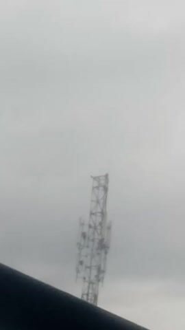 The video shows a tall cell phone tower against a cloudy, gray sky. The tower is made of a metal lattice structure and has several antennae attached to it. The tower is slightly out of focus, and the sky is blurry. In the foreground, a dark, blurry object, possibly a railing or ledge, partially obscures the view. The video appears to be shot from a low angle.