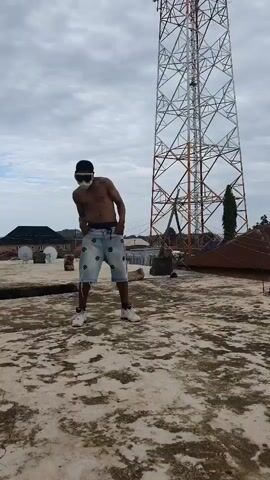 A shirtless man wearing white goggles and patterned shorts dances on a rooftop. He is positioned in front of a tall, lattice-style transmission tower under a cloudy sky. The rooftop is paved with a light-colored, textured material. In the background, several buildings with dark roofs and satellite dishes are visible.