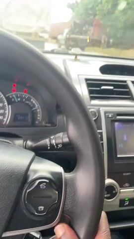 This media file provides a point-of-view perspective from inside a car, likely in Koton Karifi, Nigeria. The scene captures a moment of being in traffic, with multiple vehicles visible both ahead and to the side through the windshield.

The interior of the car is partially visible, showing the dashboard with its instruments, including a speedometer, and the car's audio system display. The audio system is playing music, with the song title "FOLK & Young Jane" visible on the screen. A car air freshener hangs from the rearview mirror. A person's hand holding a smartphone is visible in the lower right corner.

Outside the car, the weather appears overcast, suggesting it might be a cloudy day. The road is lined with other vehicles, including vans and trucks, some of which are covered with tarps. The general impression is one of congestion and waiting. The presence of multiple vans and trucks, along with the road conditions, hints at a location that might be a transportation hub or a busy route. The overall mood is one of being stuck in traffic.