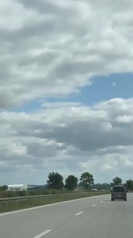 This is a vertical, low-angle shot of a highway from the perspective of a moving vehicle. The sky is filled with dramatic, grey and white clouds. On the road, a light-colored van is in the foreground, and other vehicles are visible in the distance. Trees and foliage line the side of the highway, with hints of distant windmills and buildings in the background. The road appears to be a multi-lane highway with white lane markings. The overall impression is of a journey on a cloudy day.