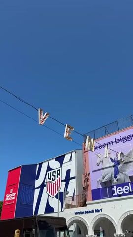A bright sunny day unfolds as the camera pans across a bustling street scene. Above, illuminated letters spelling out "VENE" are strung across wires, hinting at a larger sign. Below, a large building is adorned with a vibrant mural showcasing the USA soccer team's emblem. To the left of the mural, a red section of the building displays the "Soccer House" logo. In front of the mural, a brown UPS delivery truck is parked, its side door slightly ajar. Further left, a bright green electric truck is visible. The right side of the frame features a building with a large, colorful graphic of a person riding a white horse, with the text "dream big" above it. The bottom of this building reveals arched entrances, suggesting a commercial area. The overall atmosphere is lively and urban, bathed in the clear blue sky.