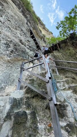 From a low angle, the video shows a rocky cliff face with a winding wooden staircase attached to it. The staircase is made of rough-hewn planks, with wooden railings and cross-bracing for support. A blue rope is tied to the lower part of the staircase. Two people are visible on the staircase. The person closest to the camera is a young woman wearing a white shirt and light-colored shorts. She appears to be pausing on the steps. Above her, a person in darker clothing is ascending the stairs. The cliff face is a light grey color, with green vegetation growing at the top. The sky is blue with some white clouds. The overall impression is of a precarious but scenic climb.