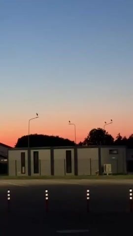A video shows a street scene at dusk. The sky is a gradient of blue and orange, transitioning from deep blue at the top to a soft orange near the horizon. In the foreground, there are several orange and white traffic cones lined up. Behind the cones, a paved area leads to a low building with a fence in front of it. Several streetlights are visible, some illuminated. To the right, there are trees and more buildings. A large truck with a trailer is parked in the middle ground, and another truck with a tanker trailer is visible further to the right. The overall impression is of a truck stop or industrial area at the end of the day.