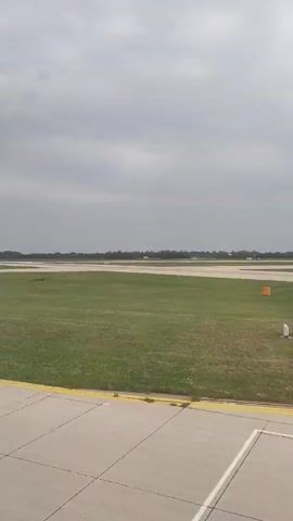 A short video clip shows an airplane taking off from a runway under a cloudy sky. The camera angle is low, possibly from another aircraft or a vehicle on the ground. The airplane is white with dark markings on its tail and appears to be ascending steeply. The surrounding environment consists of a grassy field with a paved runway marked with yellow lines. In the distance, there are trees and other airport infrastructure. The audio consists of airplane engine sounds and muffled speech.