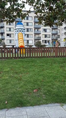 The video shows a children's playground with a large, colorful slide. A wooden fence encloses the playground area, which has a grassy lawn. In the background, a tall apartment building stands, with visible windows and balconies. A few people are present in the playground, including a child or two playing on the equipment. The overall scene suggests an urban or suburban setting. The video captures a brief moment in time within this environment.