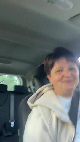 The video shows a woman with short, dark hair and a light-colored jacket sitting in the driver's seat of a car. She is looking to the right and appears to be speaking. The interior of the car is visible, including the headrests of the seats behind her. The lighting suggests it is daytime. The woman is wearing a seatbelt.