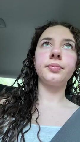 The video shows two young people riding in a car. First a young woman with long, dark curly hair smiles at the camera. Then, the camera shifts to show a young man wearing sunglasses and leaning back in his seat. The car's interior is visible, including the gray ceiling, black leather headrest, and window. Trees can be seen passing outside the car window. Music is playing in the background.