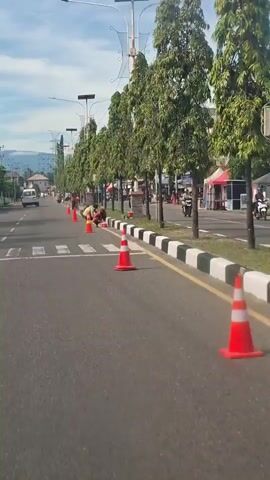 The video shows a street scene with several people working along the side of the road. Orange traffic cones are placed to mark a work area. Two people are crouched down near the curb, possibly painting or cleaning it. Another person stands and appears to be directing them or working with them. Further down the road, more people are visible, also appearing to be engaged in some form of road maintenance or cleaning. Trees line the sidewalk, and buildings are visible in the background. Traffic, including cars and motorcycles, moves along the road. The weather appears to be clear and sunny.
