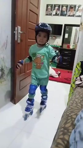 A young child wearing a helmet, wrist guards, and knee pads is standing in a room wearing roller skates. The child has dark hair and brown eyes and is wearing a green pajama set with a cartoon dinosaur chef design. The child is standing on a light-colored tiled floor with a door visible to their left. Behind the child is a bed with a patterned cover, a rug, and a dresser with a television on top. There are also framed portraits on the wall above the dresser. The child looks directly at the camera. The camera moves closer to the child during the short video.