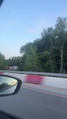 This is a first-person perspective video taken from inside a car, moving along a road. The sky is a clear blue with some wispy clouds, and it appears to be late afternoon or early evening, as evidenced by the soft lighting. Trees and foliage line the side of the road. In the foreground, a side-view mirror is visible, reflecting the passing scenery. Further down the road, a barrier with red and white markings is present, along with traffic lights and streetlights. Other vehicles can be seen on the road ahead, moving in the same direction. The video captures the smooth motion of the car traveling on what seems to be a highway or a major road.