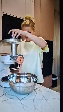 The video shows a woman with blonde hair in a bun, wearing a light yellow t-shirt, standing in a kitchen. She is operating a meat grinder attached to a white stand mixer. The meat grinder is producing ground meat, which is falling into a large silver bowl placed in front of it. The woman is holding a black cylindrical object in her right hand and appears to be feeding meat into the grinder with her left hand. The kitchen has modern cabinetry, marble countertops, and a dark-colored range hood above the grinder. A trash can is visible in the background. The woman is looking down at the meat grinder and seems to be engaged in the process of grinding meat.