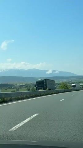 The video shows a bright, sunny day on a highway in a mountainous region, likely near Staro Selo, Bulgaria. The sky is clear blue with scattered white clouds. The camera appears to be in a vehicle moving forward, passing other vehicles.

Initially, a large green semi-truck is visible on the left side of the road, behind a metal guardrail. The truck's trailer has the text "Heiligenmayr" and "International" printed on it. Mountains with patches of snow are visible in the distant background, along with green valleys and fields.

As the camera moves, it passes the green truck, which seems to be either parked or moving slowly on the shoulder. Another white semi-truck is then briefly seen on the right side of the road, also seemingly stationary or moving slowly, before the view opens up to the expansive mountain landscape and green fields in the valley below. A metal guardrail runs along the side of the road throughout the video.