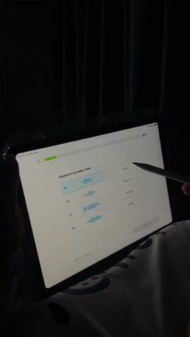 A person is holding a tablet in portrait mode. The tablet screen displays a language learning application. The app has a title in Russian that translates to "Match the pairs of words." Below the title are several interactive elements. On the left side of the screen, there are three rows, each containing a green button labeled with a word in Russian and a visual representation of a sound wave. On the right side of the screen, there are four green buttons with Russian words: "снова" (again), "таблицы" (tables), "помощь" (help), and "масло" (butter). The user is interacting with the tablet using a stylus, and it appears they are trying to match words by selecting them. The background is dark, with vertical lines suggesting curtains or blinds. The lighting is dim, with the tablet screen being the primary light source. The user's hands and parts of their body are visible, resting on a patterned surface, possibly a blanket or pillow with a smiling face design. The time stamp on the tablet indicates a few seconds have passed, and the progress bar at the top of the screen shows some progress has been made in the learning exercise. The user selects and then clicks on the sound wave visualization associated with the word "снова" (again).