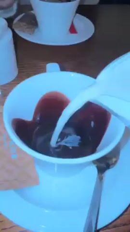 The video shows someone pouring milk into a white cup filled with coffee. There's also a sugar packet with the words "CAFE RICH" on the side of the cup, and a small dessert on a saucer behind the cup. The table appears to be made of dark wood.