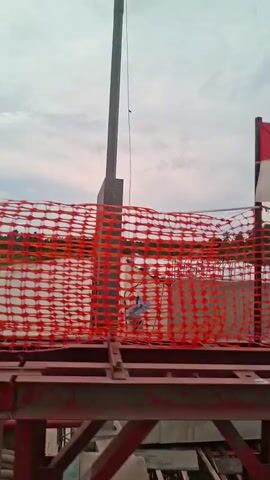 The video shows a view from a moving vehicle on a bridge. There is a red safety net in the foreground and a large metal structure on either side of the frame. The bridge appears to be under construction, with a temporary fence visible in some shots. A body of water, possibly a river, is visible below the bridge, and the surrounding area is lush with vegetation. The sky is overcast.