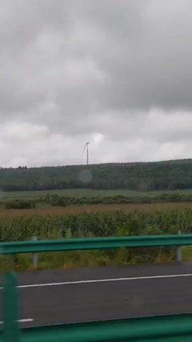 The video captures a scene from inside a moving vehicle, looking out at a rural landscape under a cloudy sky. In the distance, atop a hill covered with trees, a single wind turbine is visible. Its blades are slowly rotating. In the foreground, there is a green highway barrier and a paved road. Between the barrier and the hill, there are fields of crops, possibly corn, and some bushes. The overall impression is of a natural, open environment.