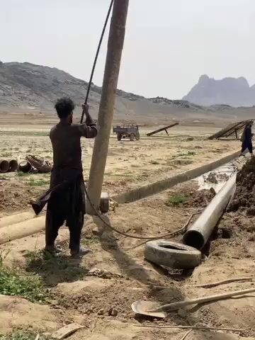 The video captures a well-drilling operation in a rural, arid landscape in Nowruzi, Afghanistan, under bright daylight.

In the foreground, a man in dark traditional clothing stands next to a tall, thick wooden pole, holding onto a black hose or rope that runs up the pole. He is actively involved in operating a drilling rig. To his right, a partially excavated trench contains water and large pipes, with a shovel and a discarded tire visible on the ground nearby.

The camera pans right, revealing the full drilling setup. A large, green, spoked wheel rotates rapidly, driven by a belt connected to an engine assembly. This machinery is responsible for boring into the earth. Another man can be seen walking in the background further to the right. The engine is positioned near a blue barrel.

The background features rugged, rocky mountains under a clear sky. In the mid-ground, several solar panels are angled towards the sun, likely used for power or irrigation. Beyond the immediate dusty work area, a field with green crops is visible, contrasting with the predominantly dry and barren surroundings. The scene depicts a focused effort to access water, highlighting the use of mechanical equipment in a remote and resource-challenged environment.