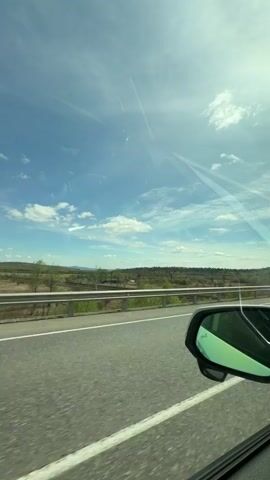 The video shows a view from inside a car driving on a highway. The sky is blue with scattered white clouds. The sun is shining. There is a green landscape with hills in the distance, visible over a metal guardrail lining the road. The car's side mirror is visible in the foreground. The road is gray asphalt with painted white lines.
