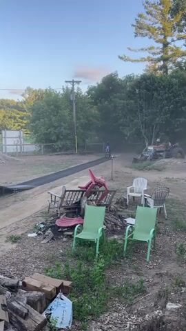 A dirt bike rider approaches a ramp, launches into the air, and performs a trick before landing on an inflatable airbag. The scene takes place outdoors in a rural setting with trees and a blue sky. Several outdoor chairs and piles of wood are scattered in the foreground.
