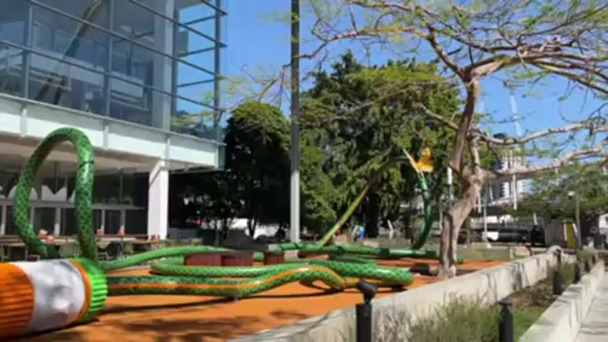 A low-angle shot depicts a playful public art installation resembling a large green garden hose with orange stripes. The hose stretches across a brown, mulch-covered area near a modern glass building and green trees. The hose ends with a white and orange nozzle, positioned prominently in the foreground. Behind the hose is an outdoor restaurant seating area. In the distance, a street with parked vehicles and a construction crane is visible. A person with a cleaning cart walks into the frame behind the hose. The sky is clear and blue, indicating a sunny day.