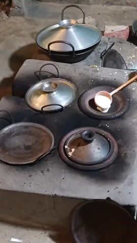 The video shows a close-up shot of a traditional clay stove with multiple cooking surfaces. There are several pots and pans on the stove, some covered and some open. In the foreground, a ladle is pouring a white batter into a round, shallow pan. The batter is spreading out evenly. The stove is blackened with soot, indicating it has been used for cooking. The background is dimly lit and appears to be an outdoor or rustic setting. There are no people fully visible in the frame, but a hand is visible holding the ladle. The overall scene depicts food preparation using a traditional cooking method.