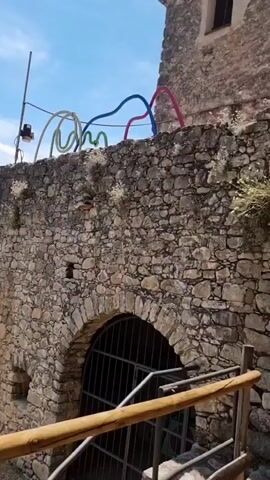 The video pans across an old stone building with arched doorways. The walls are constructed of rough, irregular stones. In the first shot, colorful, curved structures are visible above the stone wall. In the second shot, the camera moves upwards to show the side of a building with two small windows. The third shot shows an arched doorway with a dark door. The fourth shot shows the corner of a stone wall. The fifth shot shows a stone building with a rounded section and windows, surrounded by trees. The lighting suggests it is daytime.