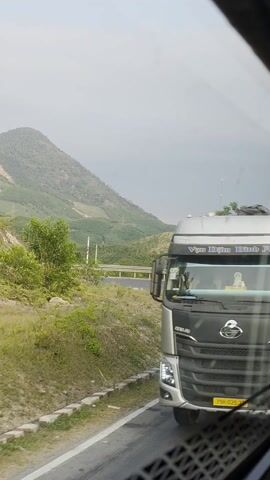 The video shows a view from a moving vehicle, possibly a car or bus, driving on a winding road in a hilly area. Initially, the view is partially obstructed by the side of a large truck, but the scene quickly shifts to a clear view of the surrounding landscape. The landscape is characterized by green hills and mountains under a cloudy sky. The road has a guardrail along the side, and there is a road marker visible. The vegetation appears lush, suggesting a temperate or tropical climate. Some deforestation is visible on the hillside in the background.