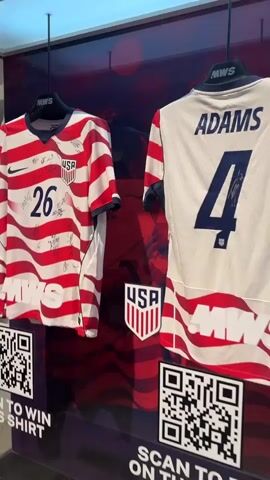 A display case contains two US Soccer jerseys. The jersey on the left is red and white striped with the number 26 on the back and is signed by multiple players. The jersey on the right is white with red and blue stripes on the sleeves and collar, the name ADAMS and the number 4 on the back, and is also signed. Both jerseys have QR codes below them. The left QR code says "SCAN TO WIN THIS SHIRT" and the right QR code says "SCAN TO BID ON THIS SHIRT". Above the jerseys, the text "OWN A PIECE OF SOCCER HISTORY" is visible. The background behind the jerseys is a blurred image of a US flag. The display case is made of glass and metal and is placed on a concrete floor.