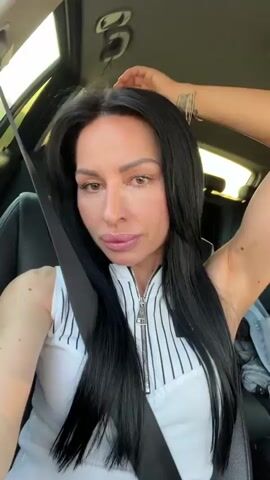A woman with long, straight dark hair, wearing a white sleeveless top with black vertical stripes and a zippered front, is seen in a car. She is in the driver's seat, secured by a seatbelt across her chest. Her arm is initially raised, revealing a tattoo on her forearm, and her fingernails are painted pink.

The woman smiles and poses for the camera, which appears to be held by herself, indicating a selfie video. She frequently looks into the lens, showing a cheerful and engaged expression. Her makeup is visible, including pink lip gloss.

Through the car window, a bright, sunny daytime scene unfolds, suggesting the vehicle is in motion, likely on a road flanked by trees and fields. The interior of the car is dark, providing a contrast to the bright exterior. At various points, she adjusts her hair and continues to smile and look at the camera. A small, unidentifiable logo or text is visible on a black patch on her top. Background audio includes music or other sounds, but no discernible speech from the woman.