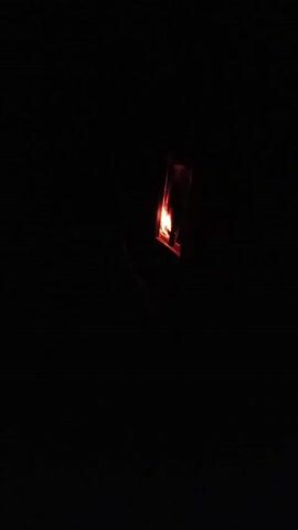 In extreme darkness, the flame of a lighter is visible. The lighter is silver in color. The flame is orange.