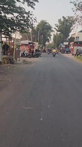 A street scene is shown at dusk. Motorbikes are parked and being ridden on the road. There are several small stalls and shops lining the street, selling various goods. The sky is a pale orange and the trees are casting long shadows. The overall atmosphere appears to be that of a busy marketplace or a residential area with local businesses.