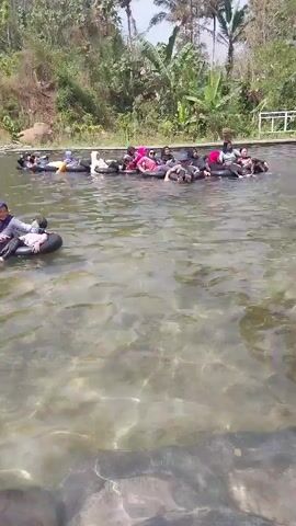 A group of people are floating on inner tubes in a clear body of water. The water is shallow enough to see the rocky bottom. The background is lush green with trees and vegetation. The people are clustered together, some talking and laughing. One person is on an inner tube closer to the camera, wearing a hijab and holding a child. There are small slides visible in the background. The sun is shining, indicating it is daytime. The overall atmosphere appears relaxed and recreational.