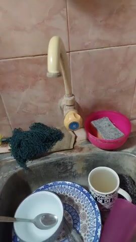 A hand turns a faucet handle, and water begins to flow from the faucet. The faucet is beige and is mounted on a tiled wall. To the left of the faucet is a pile of dark green netting. To the right of the faucet is a pink bowl containing a sponge. Below the faucet is a sink with dishes in it, including a white bowl with a spoon and a patterned plate. A white mug is also visible in the sink.