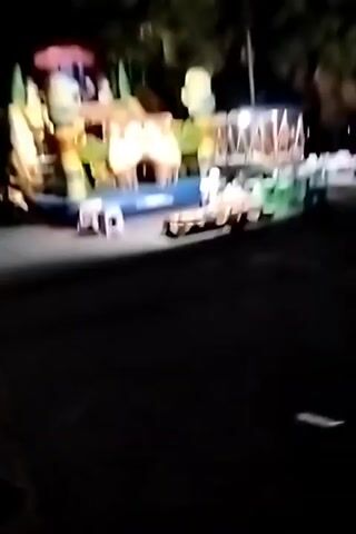 A nighttime scene shows an inflatable bounce house with various cartoon characters, possibly Minions, and a slide. In the foreground, there are several low stools and some tables and chairs. A motorcycle is parked on the right side of the bounce house. In the background, there are more tables and chairs set up under a covered area, and palm trees are visible. People are present, with one person sitting on the ground next to the bounce house. The lighting suggests it is dark, and the scene appears to be an outdoor gathering or event.