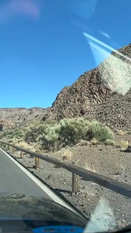 The video shows a clear blue sky over a mountainous, arid landscape. In the foreground, there is a wooden barrier with yellow markings along a paved road. The road curves into the distance, where it appears to wind through hills. Two white cars are visible driving on the road. Sparse, dry vegetation, including bushes and dead branches, dots the landscape. The mountains are rocky and rugged, with varying shades of brown and gray. The overall impression is of a remote, undeveloped, and sunny environment.