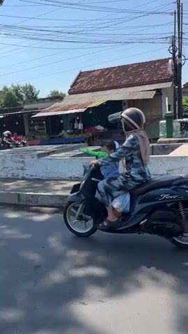 A woman wearing a hijab and a blue and white patterned dress rides a dark-colored scooter on a paved road. She is wearing a helmet. A young child, wearing a blue shirt and possibly a head covering, is seated in front of her on the scooter. The child is holding a green object. The woman is holding a clear plastic bag with something inside. The background shows buildings, including one with a red tile roof, and overhead power lines. The scene appears to be a street in a town or village on a sunny day.