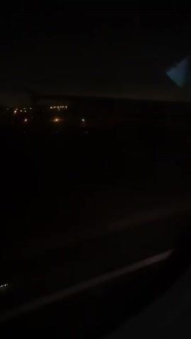 The video shows a nighttime view from an airplane window. The ground below is dark, with scattered lights visible, suggesting a city or town. The lights appear to be from buildings and possibly streetlights. The wing of the airplane is partially visible in the frame. The scene is dimly lit, with only the lights on the ground and the wing providing illumination.
