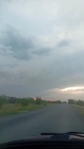 The video shows a first-person perspective of someone driving on a paved road. The road is bordered by green grassy areas and scattered trees and bushes. The sky is overcast with gray clouds, and there is a faint glow of sunset visible on the horizon, suggesting it is either dawn or dusk. The overall impression is of a rural landscape.