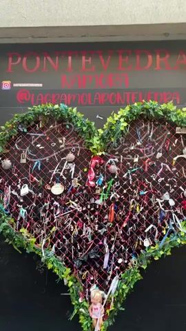 This is a vertical, shaky video of a large heart-shaped structure adorned with ivy on its edges. The heart is filled with a red mesh net on which numerous padlocks and ribbons are attached. The padlocks vary in size and color, and the ribbons are in a multitude of colors, many tied around the padlocks. In the upper portion of the video, there are some words visible in large, stylized lettering on a dark background. The words appear to be "PONTEVEDRA" and "NAMORA". There is also a social media handle "@lagraciamolapontevedra" visible. At the bottom of the heart-shaped structure, a small teddy bear is attached. The lighting suggests an outdoor environment, possibly daytime. The video is blurry and shaky, making it difficult to discern finer details.
