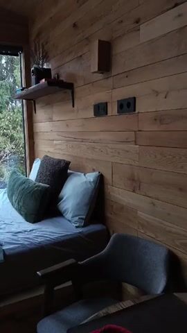 The video shows a modern cabin interior, primarily focusing on sleeping areas. The walls are covered with light-colored wood planks. There are two beds, one with gray linens and green and brown pillows, and the other, near a window, featuring brown robes and a white towel. In another section, a bed is adorned with a teddy bear and a plaid blanket. A black wood-burning stove stands against a wall decorated with gray stone. Large windows provide views of a snowy forest outside. There is a gray chair in the foreground and a shelf with objects on the wall.